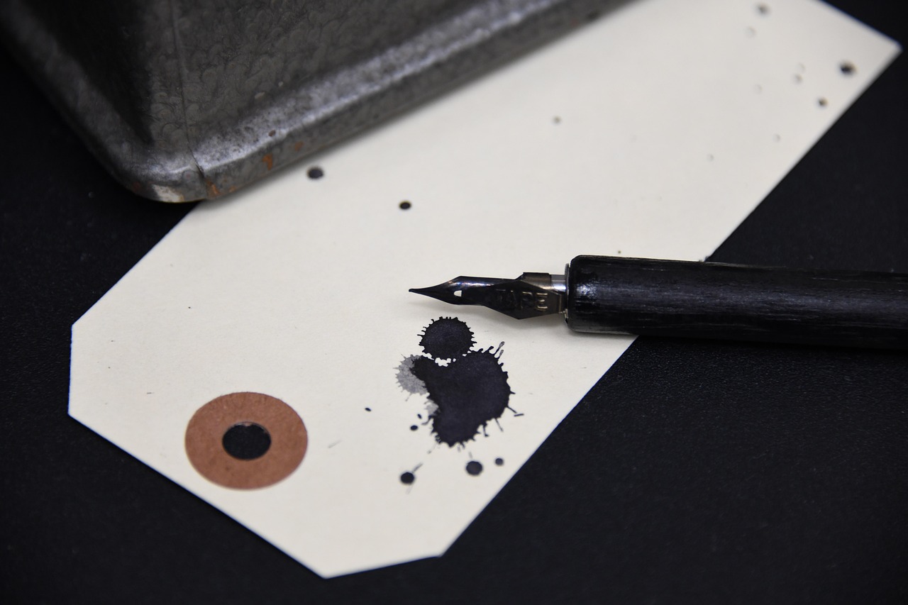 Dip pen nib beside an ink blot on a paper tag
